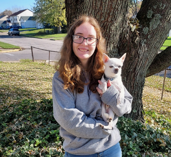Crystal, professional dog groomer in Columbus Ohio, holding a freshly groomed white Chihuahua in a cozy sweatshirt outdoors near a tree.